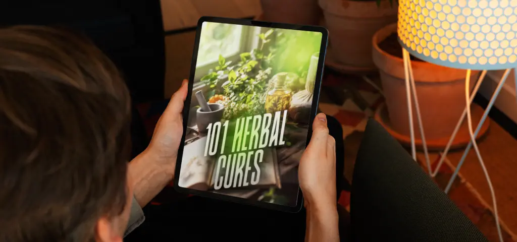 Person holding a tablet displaying "101 Herbal Cures" ebook cover with herbs, mortar and pestle on a table